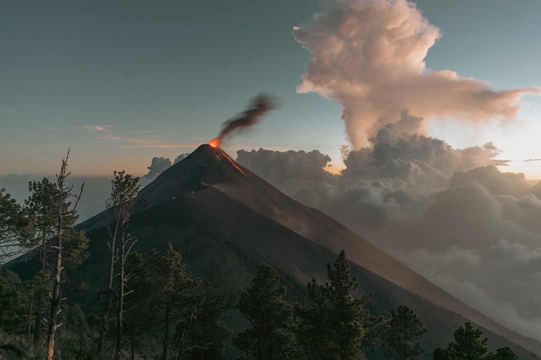 A black volcano standing tall against a dusky sky, lava is flowing down it's slopes and black smoke is billowing from the top. Volcan de Fuego, Acatenango volcano overnight hike, Guatemala