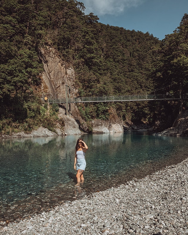 A girl standing in a blue river. Behind her is a swing bridge with people crossing. Blue Pools, Mt Aspiring National Park.