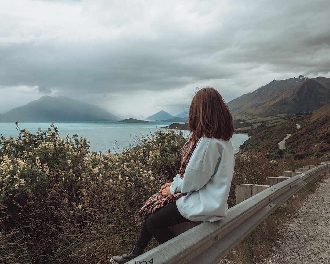 A girl sitting on the side of the road looking out over a lake. At the top of the lake are mountains under a grey gloomy sky. Queenstown to Glenorchy