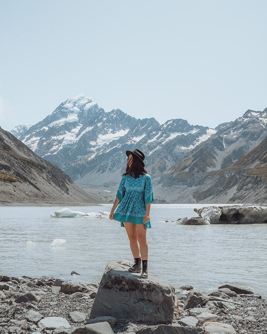 A woman wearing a blue dress and a black hat stands on a rocky terrain, gazing towards snow-capped mountains under a clear blue sky. A glacial lake with floating ice chunks lies in front of the rugged mountain landscape. Hooker Valley Track.