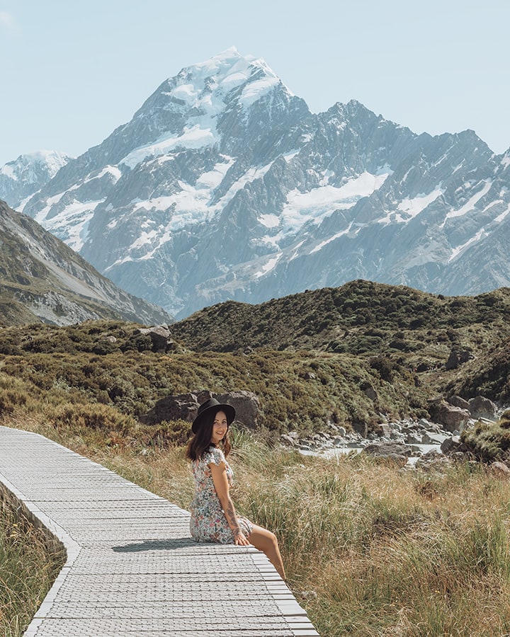 A girl sitting on a wooden walkway over tall grass. Behind her is a tall mountain covered in snow. Hooker Valley Track, New Zealand South Island Itinerary