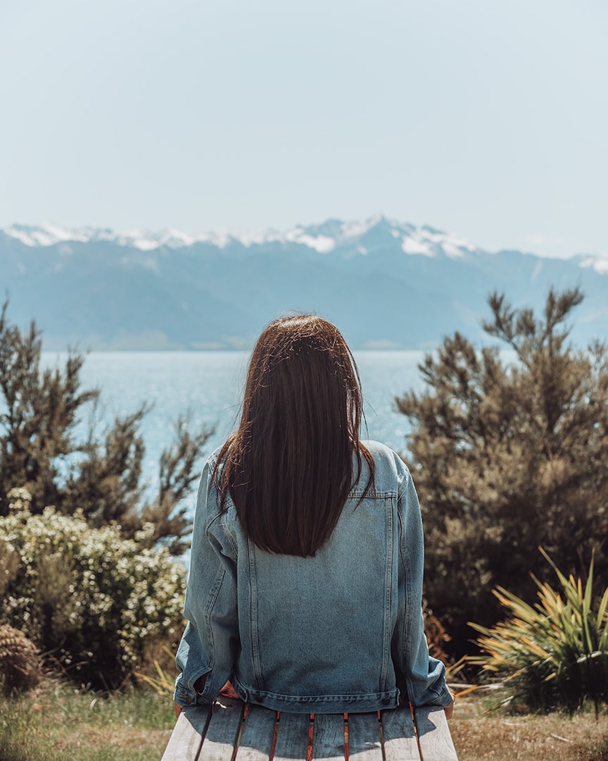 A girl sitting on a bench looking out over a blue lake. Behind the lake are snowy mountains against a blue sky. Lake Hawea, New Zealand South Island