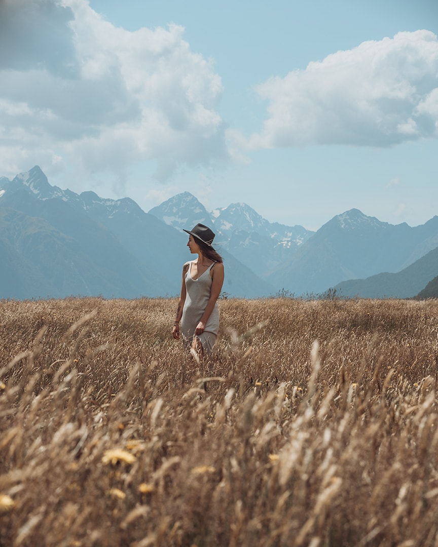 A girl standing in a field of tall grass, behind her are snowy mountains. Eglinton Valley, Milford Sound.