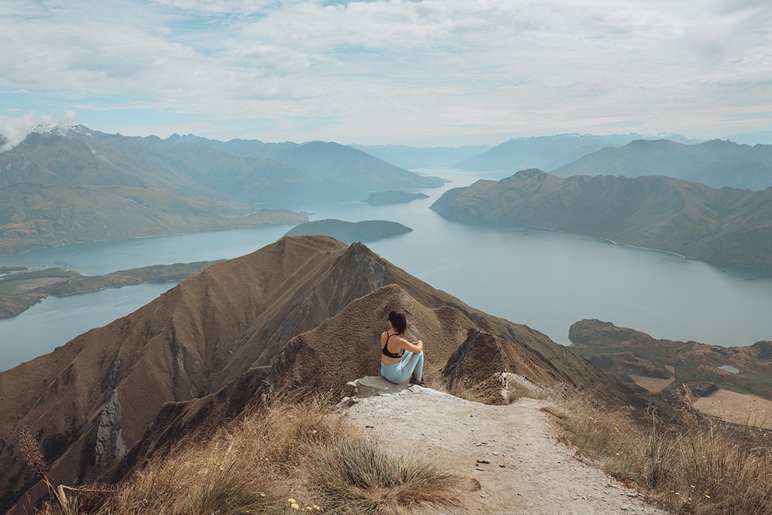 A girl sitting at the top of a mountain. She is looking out over other mountains and at a glacial lake below. Roy's Peak, Wanaka. NZ Roadtrip