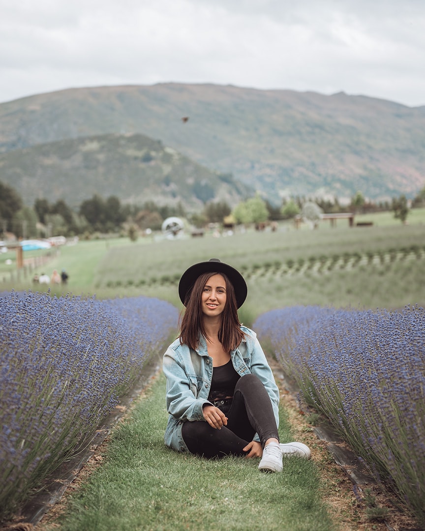 A girl sitting in between rows of purple lavender. Behind her is a green mountain. Wanaka Lavender Farm