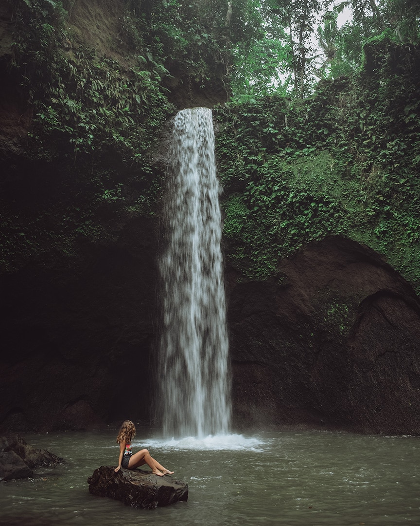 A girl sitting on a rock in water. In front of her is a waterfall.