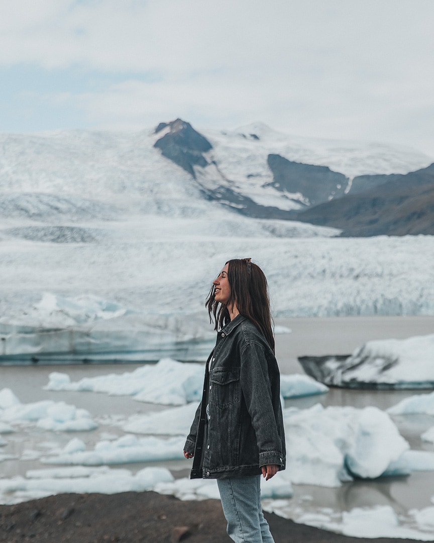 A woman stands in profile against a backdrop of a glacier with floating icebergs in the water in front. She wears a dark denim jacket and jeans, with her hair down.