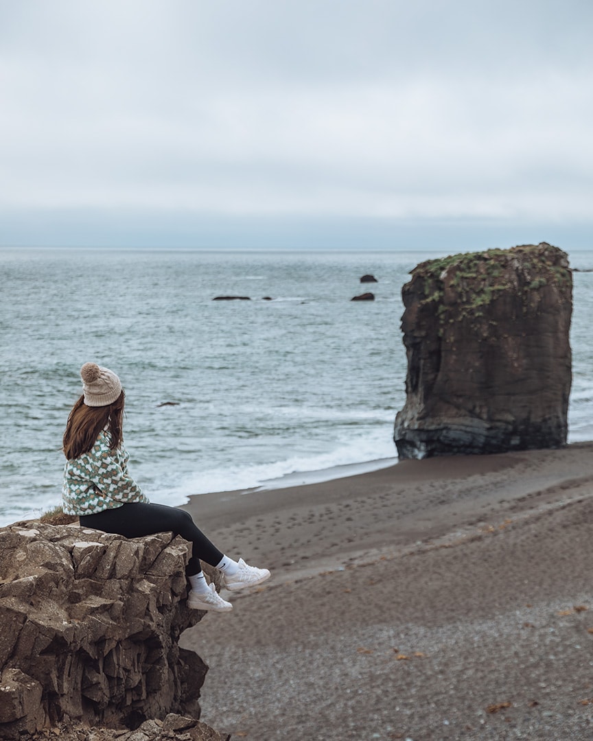 A girl wearing a fluffy white and green jacket and a cream wooly hat. She is sitting on the edge of a cliff overlooking a grey rocky beach with a tall boulder on the waters edge.