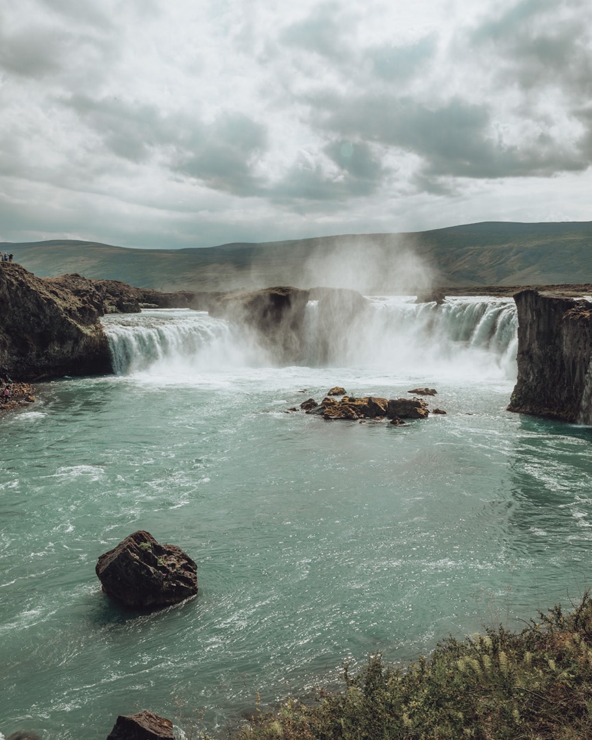 Two waterfalls next to each other poring over dark rocks sending mist up in to the air. Behind the waterfalls and green hills.