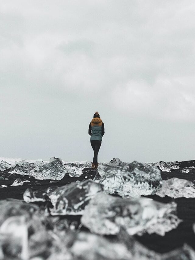 A person standing on a black sand beach. All across the sane is jagged blocks of ice that look like diamonds. Diamond Beach, Iceland
