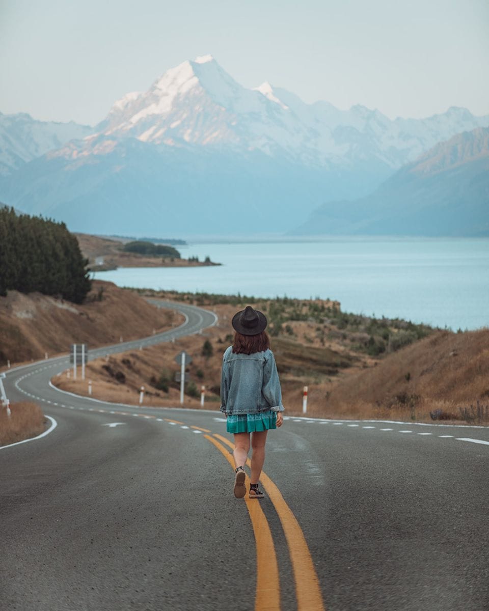 A girl walking down a road next to a turquoise lake. At the top of the lake is a tall snowy mountain. Lake Pukaki & Mount Cook, New Zealand South Island 7 day itinerary