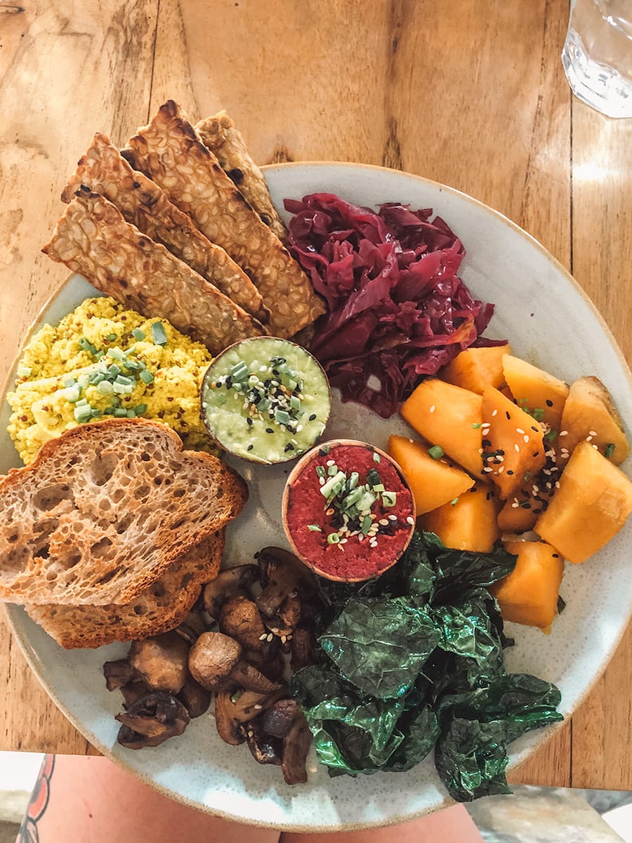 A colorful brunch plate with toast, sautéed mushrooms, kale, scrambled tofu, sliced papaya, pickled cabbage, tempeh strips, and two dips served on a wooden table.