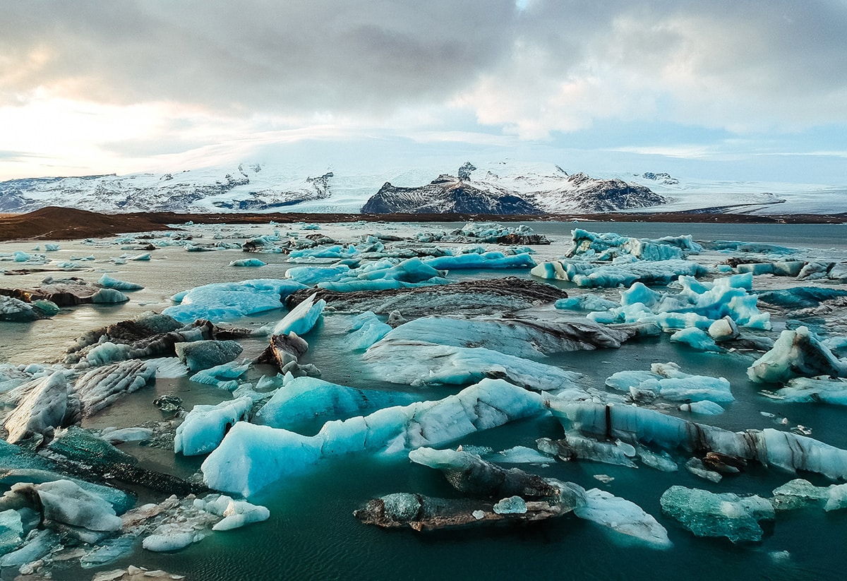A body of water filled with piercing blue icebergs. The backdrop is snow covered mountains against and cloudy sky.