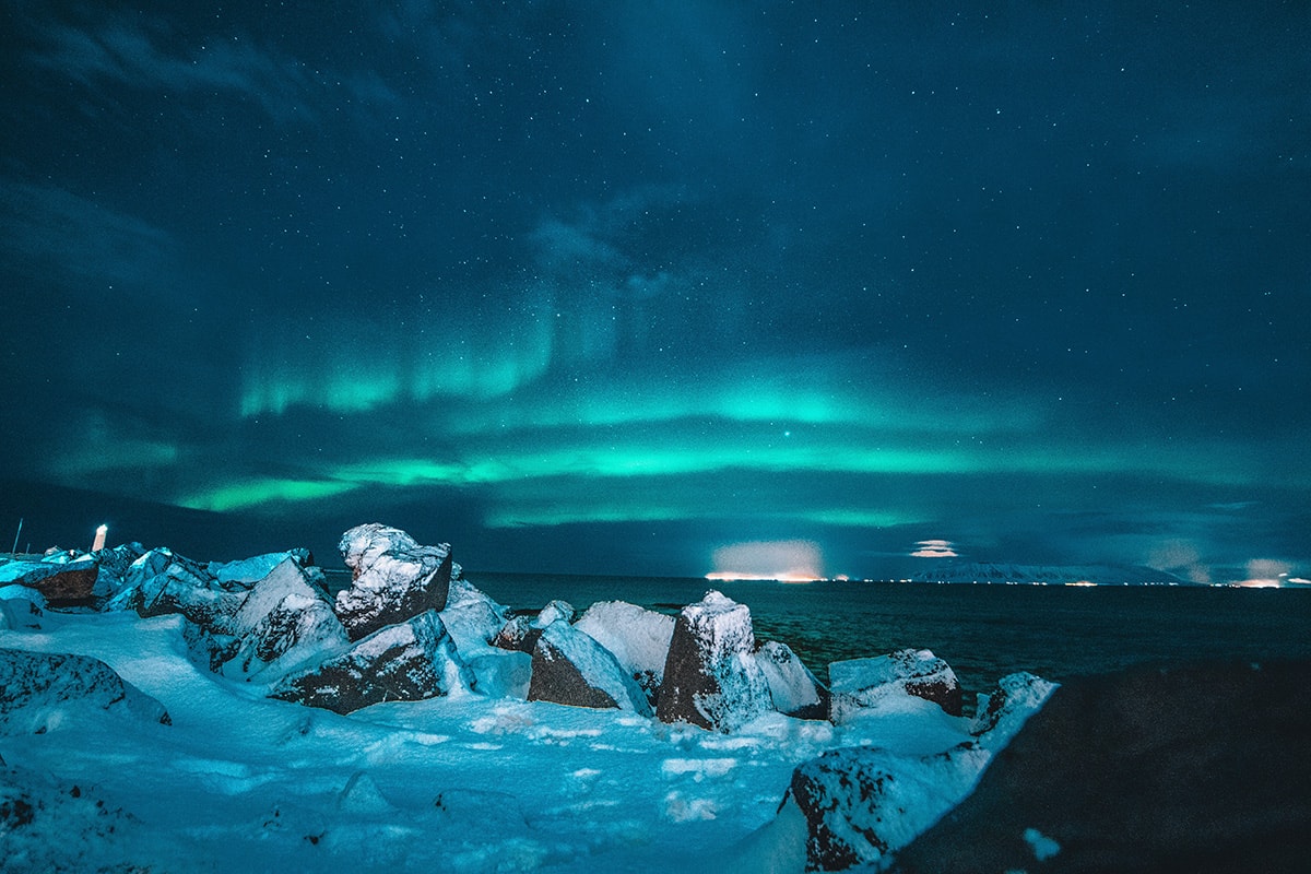 Green shimmering northern lights against a dark starry sky. Next to the ocean there are snow covered rocks.