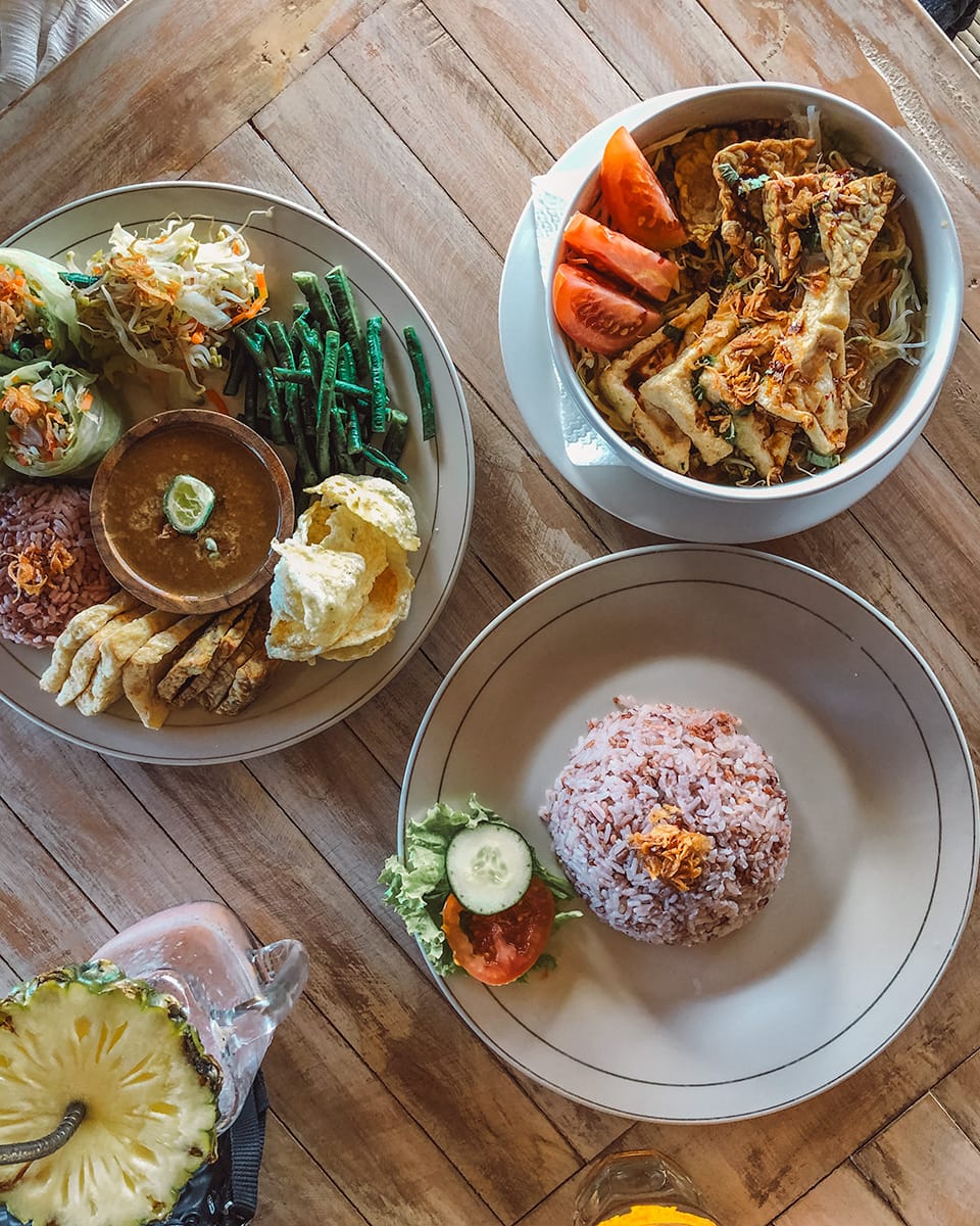 Plates of vegan food on a wooden table: noodle soup, mixed vegetables with rice, and rice with a small side salad. Vegan Soul Kitchen.
