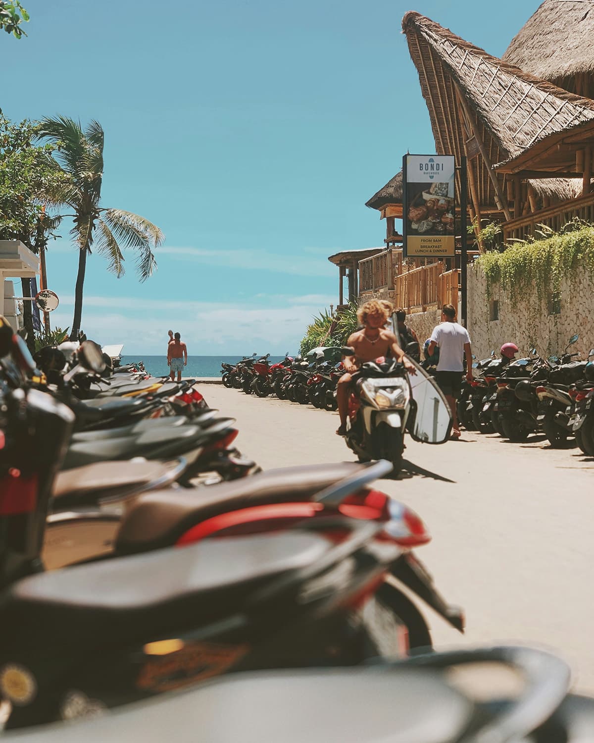 A coastal street lined with parked scooters leads down to the ocean, with a surfer riding a scooter and palm trees swaying under a bright blue sky.