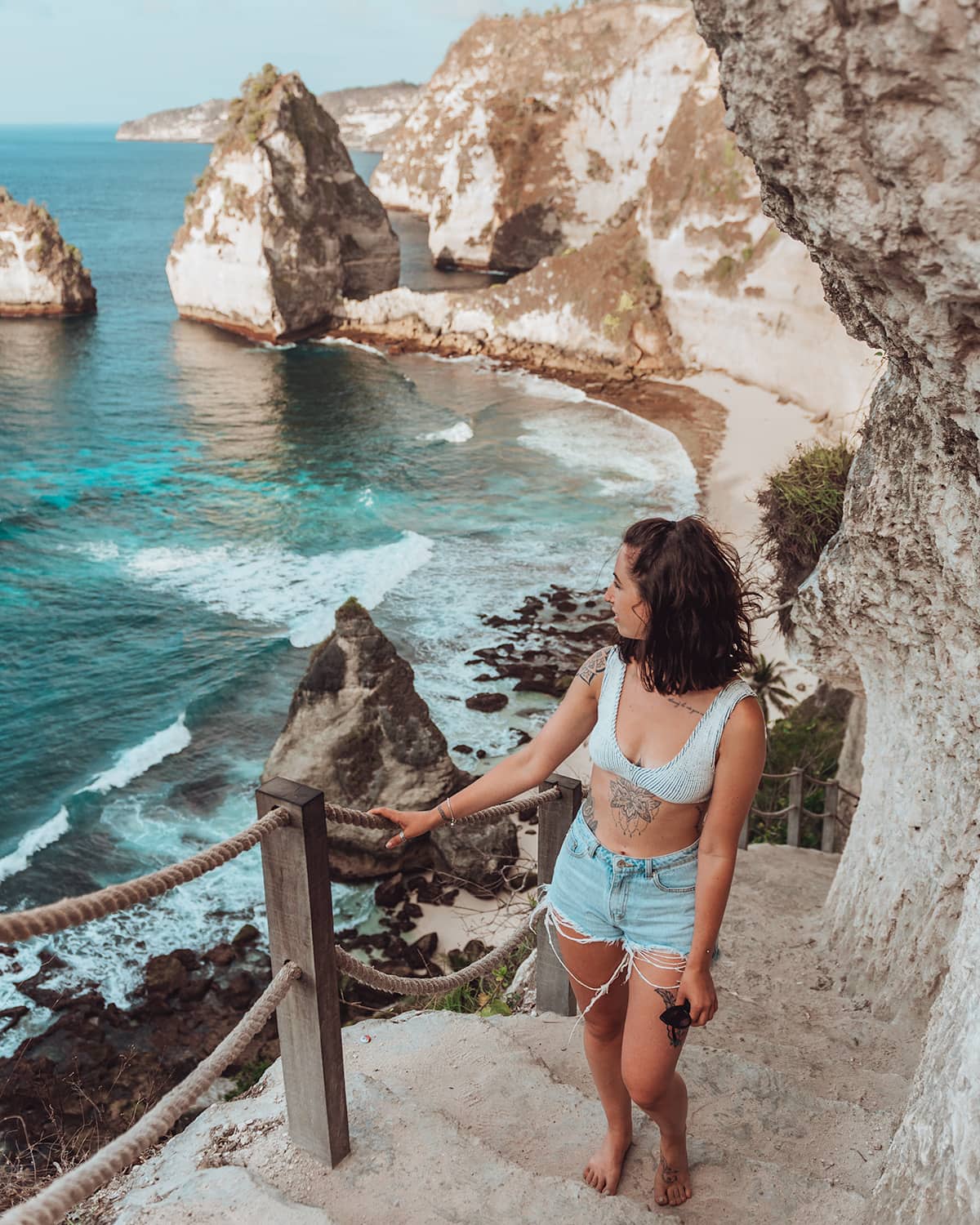 A cliffside view where the ocean meets rocky shores, with a woman looking out from a high vantage point. Diamond Beach, Nusa Penida