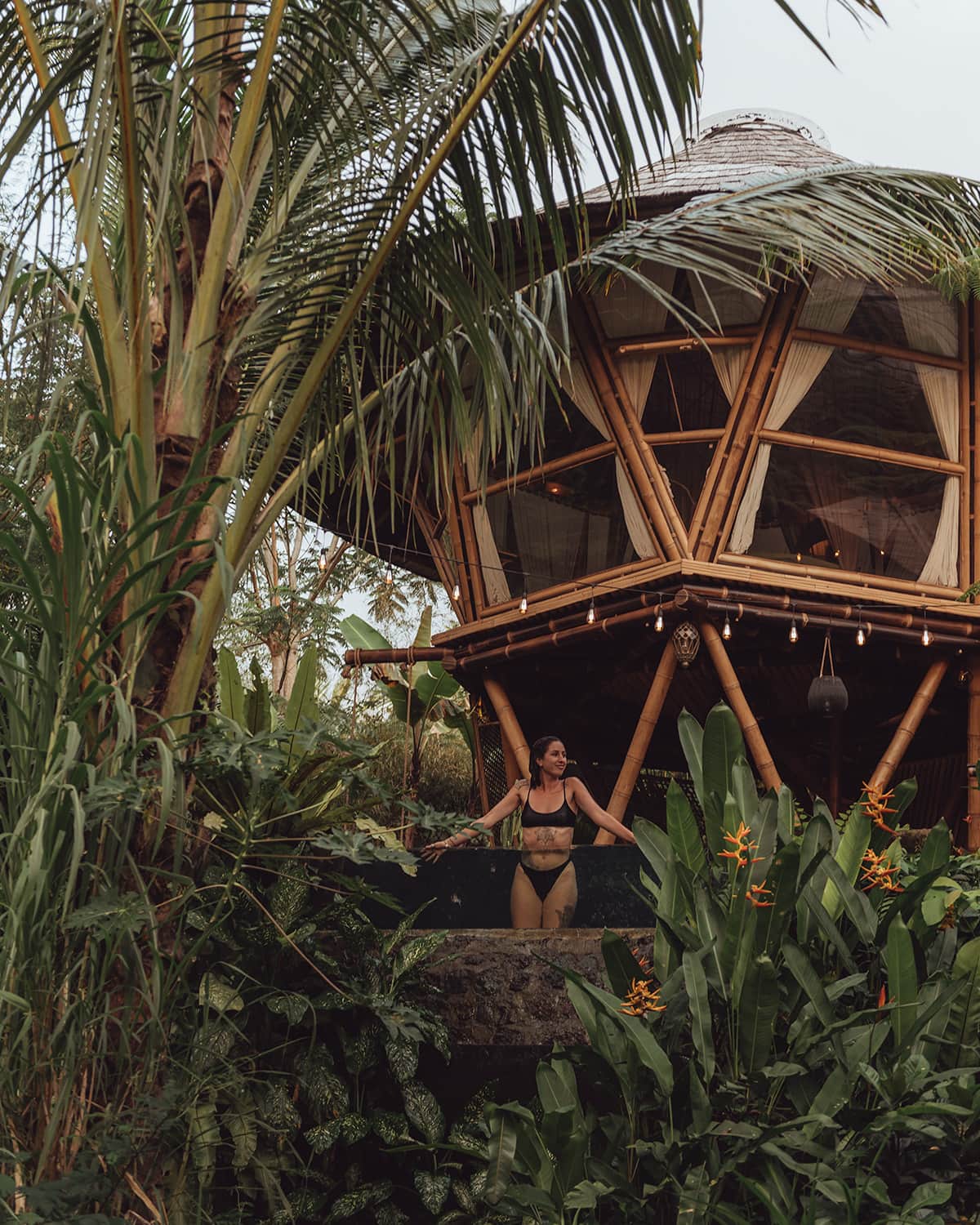 A woman stands on an elevated bamboo platform surrounded by dense tropical foliage, with a unique geometric bamboo structure in the background. Hideout, Bali