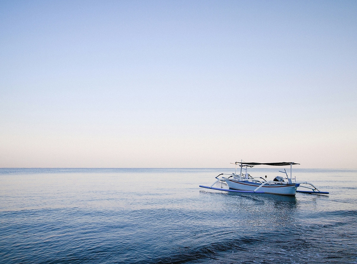 A traditional wooden outrigger boat floats on calm, glassy water under a pastel sky at dawn or dusk.