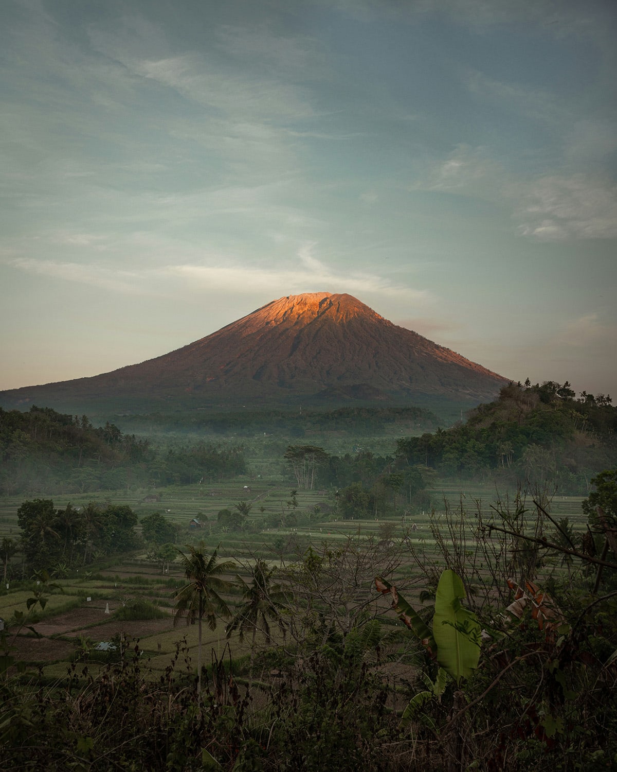 The warm glow of sunset touches the peak of Mount Agung volcano overlooking a rural landscape with palm trees and terraced fields.