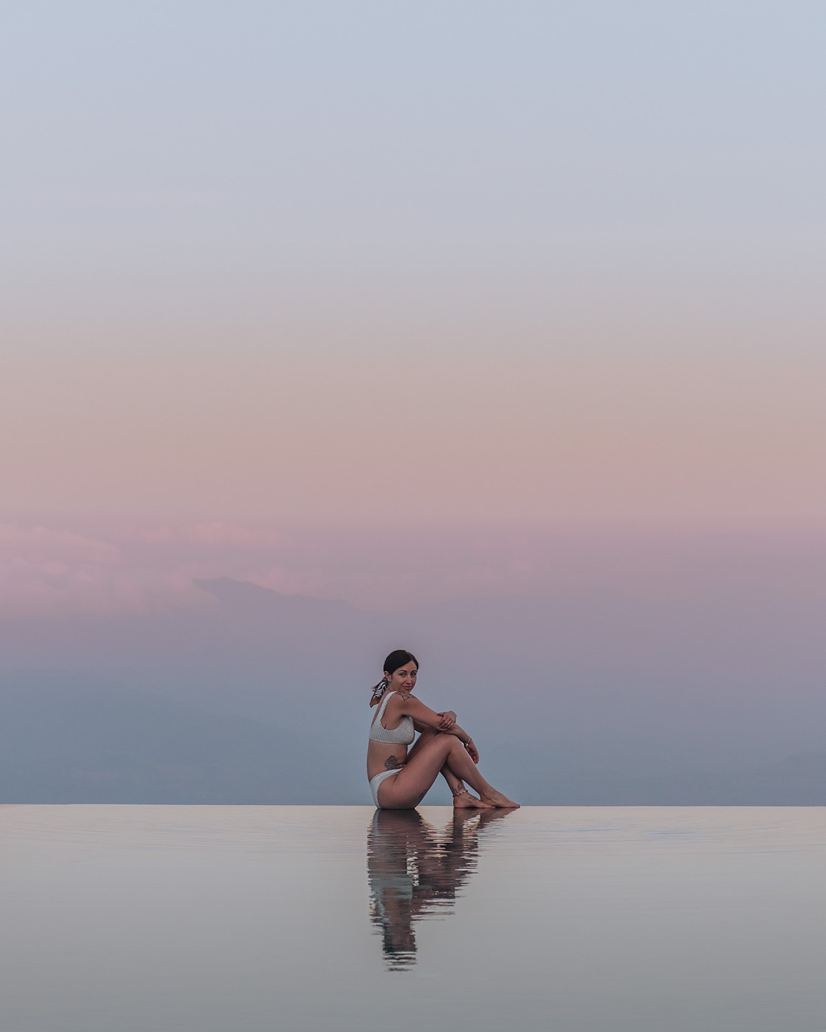 A woman sits on the edge of an infinity pool, with a backdrop of mountain silhouettes and a pastel-hued sky. Munduk Moding Plantation, one of the best places to see sunsets in Bali.