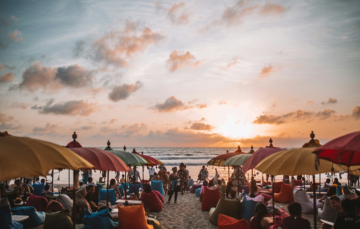 A bustling beach scene under a sunset sky with people sitting under colorful umbrellas on bean bags. La Plancha, Bali at sunset.