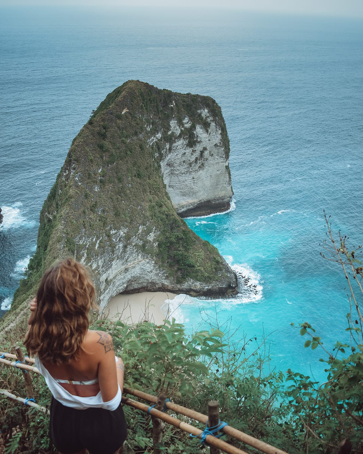 A woman gazes at a steep, lushly vegetated cliff that descends into a turquoise sea.