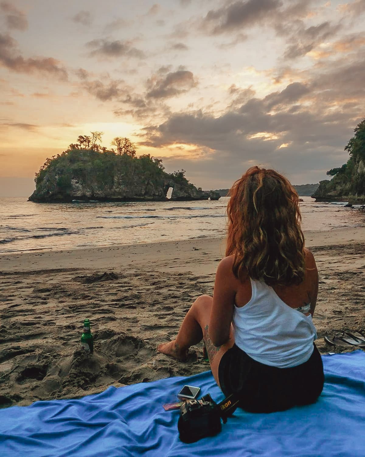 A woman sits on a beach with a golden sky facing a small, tree-covered island in the ocean.