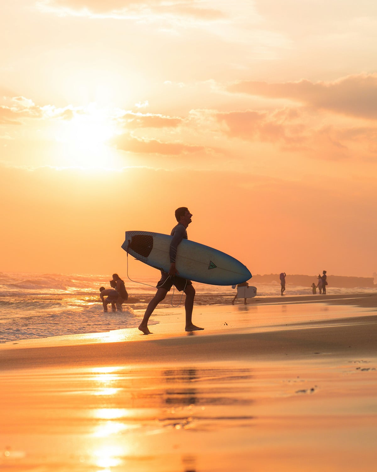 A surfer walks along a golden shore as the sun goes down with a surfboard under his arm.