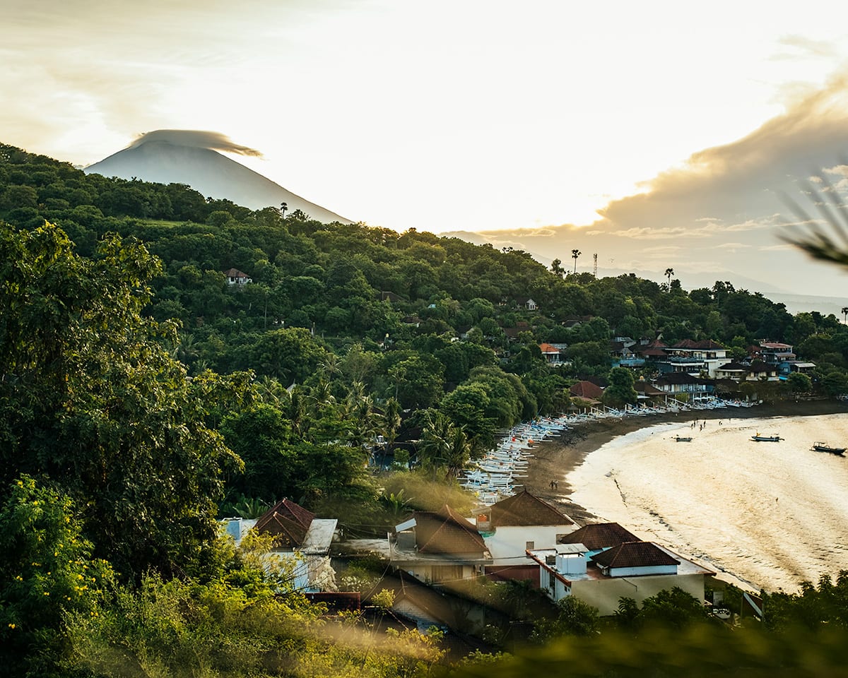 A sunset view over a bay with volcano in the distance and a tropical coastline in the foreground. Boats dot the black sand beach.