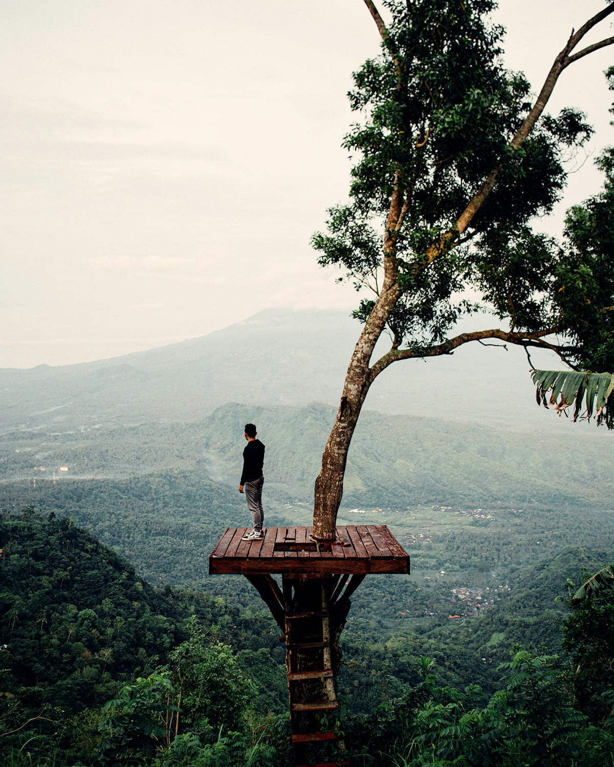 A man stands on an elevated wooden platform overlooking a vast green valley with a mountainous backdrop at golden hour in Bali.