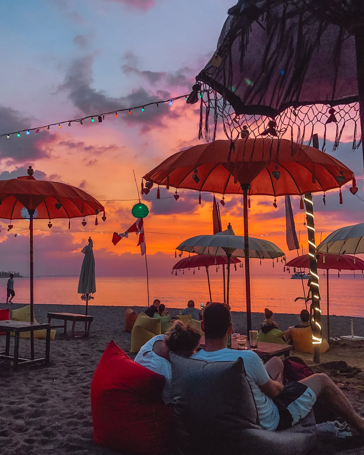 A beach with colorful umbrellas and lanterns under a pink and purple sky, with people relaxing on bean bags. Lovina Beach, one of the best places to catch sunset in Bali.