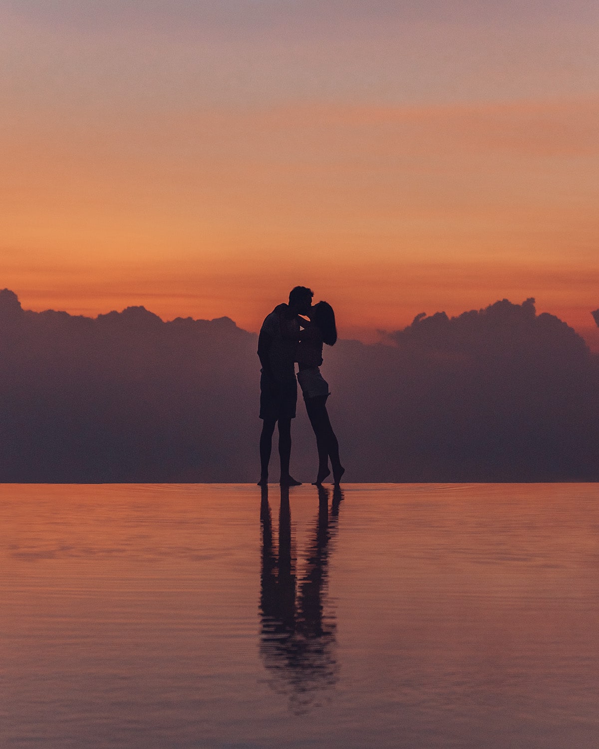 A couple hugging on a reflective water surface with the sunset creating silhouettes and a vivid orange sky.