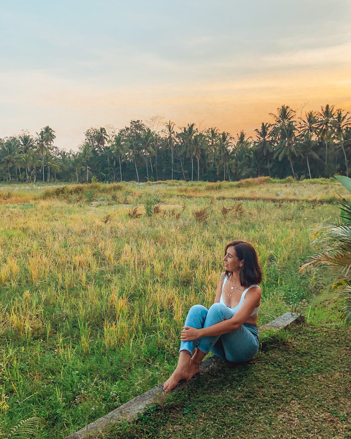 A woman in a pair of blue jeans and a white top sits on a stone wall amidst a field of tall grasses at sunset, surrounded by palm trees.