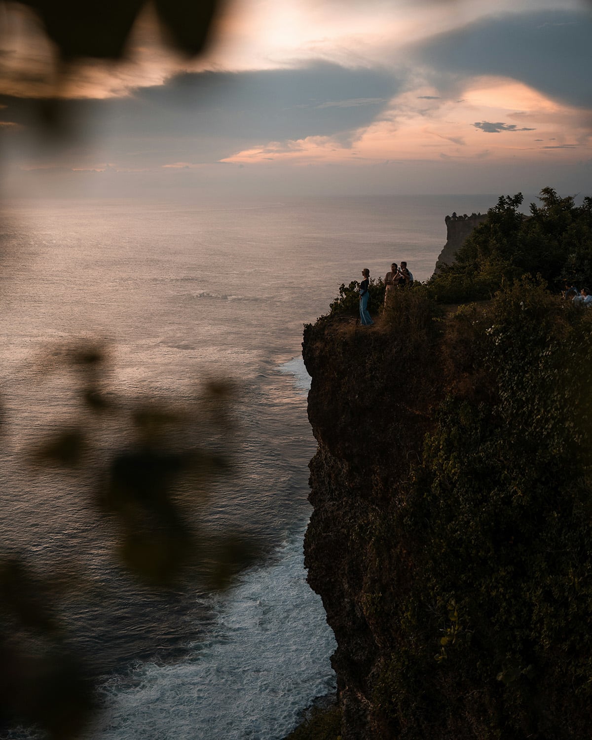 A dramatic ocean cliff at sunset with people admiring the view from the edge, framed by blurred leaves in the foreground.
