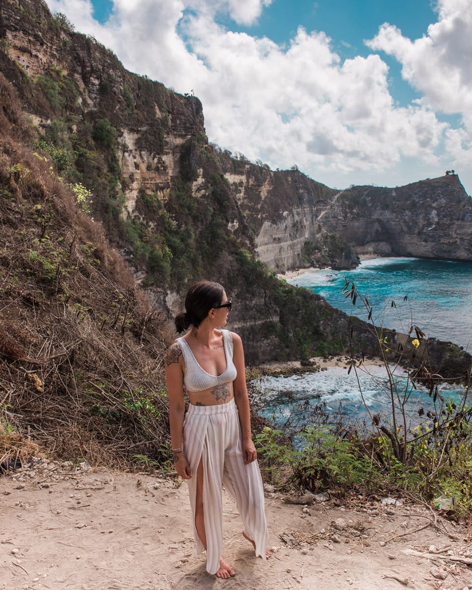 A woman on a cliff path overlooking a turquoise bay bordered by tall cliffs. Nusa Penida the last stop on 10 days in Bali itinerary.