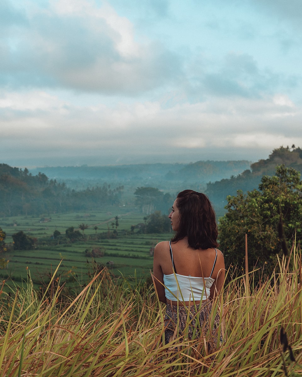 A woman, seen from behind, looks out over a verdant landscape of rolling hills and farmland. Bukit Cinta viewpoint.