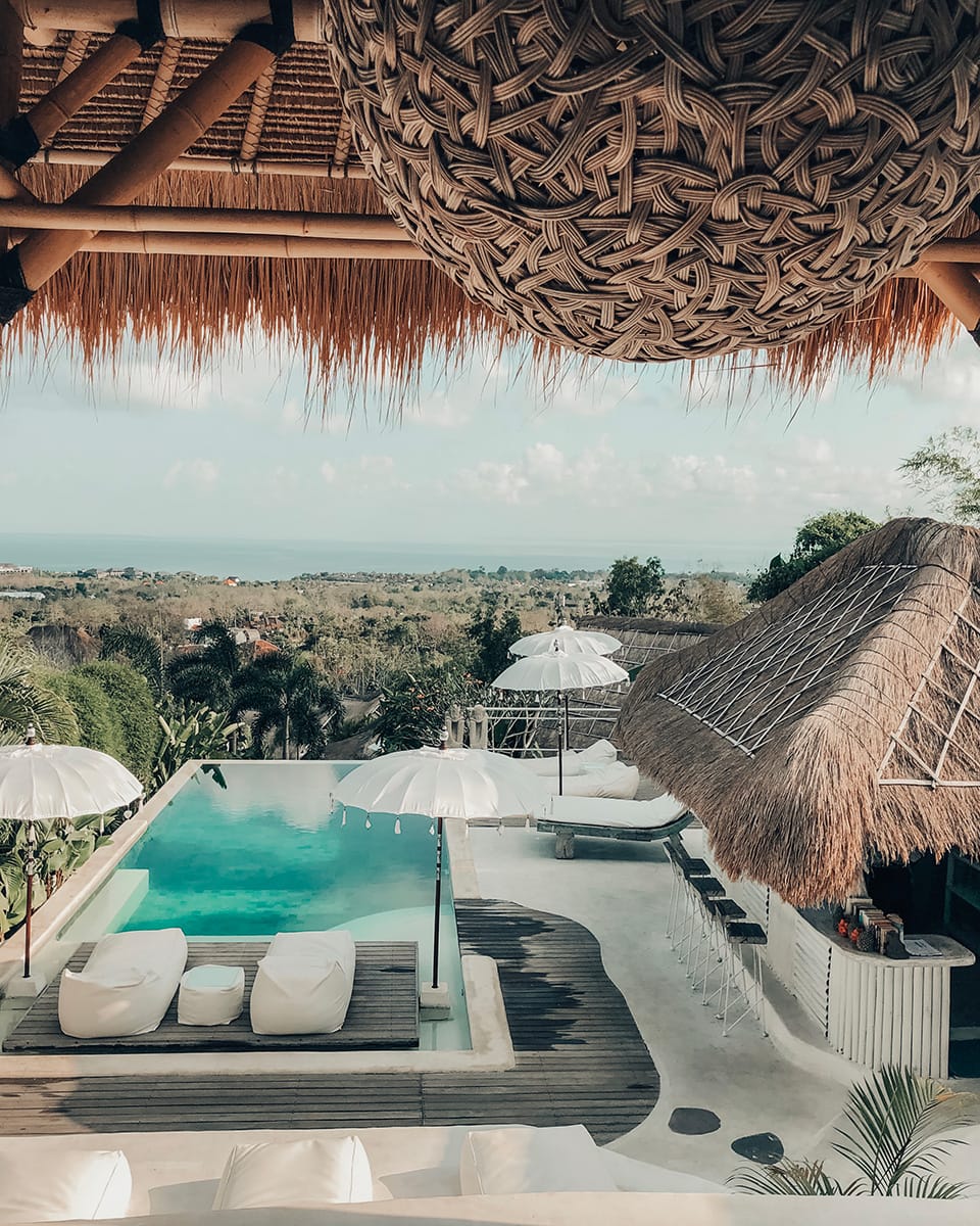 An aerial view shows an infinity pool with a woven roof structure, with a panoramic view of the landscape and ocean in the distance. Gravity Hotel, Uluwatu