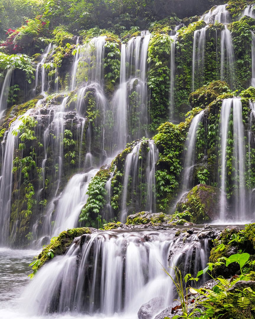 A beautiful multi-tiered waterfall cascades down a lush green cliff, with several small streams creating a curtain of water over moss-covered rocks.