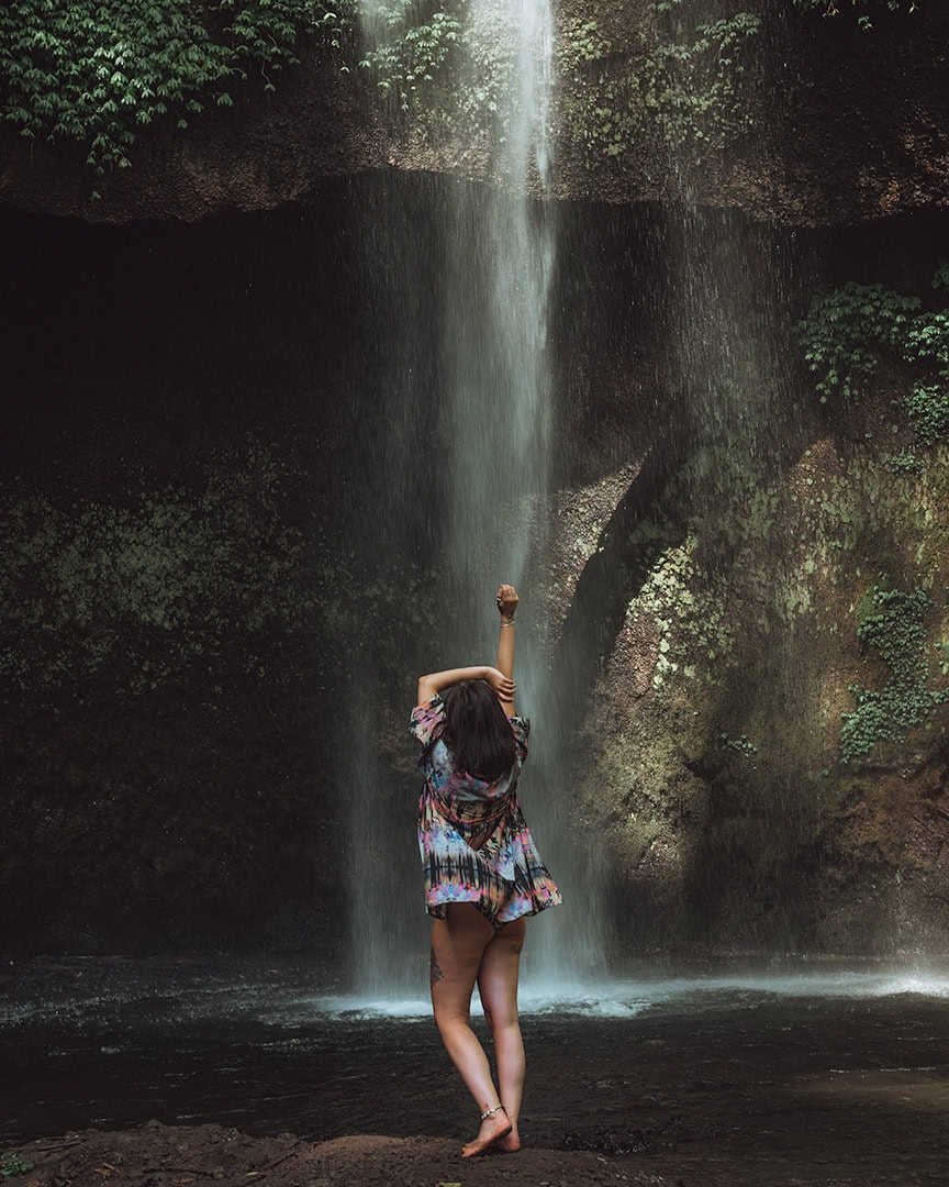A woman stands under a tall, narrow waterfall in a lush, green jungle setting. Banyu Wana Amertha Waterfall, Munduk.