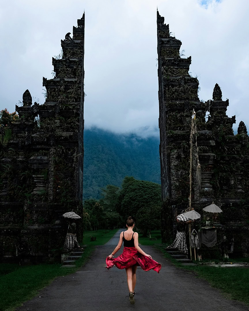 A person in a red dress walks towards a tall, ornate stone gate with mountains in the background. Handara Gate