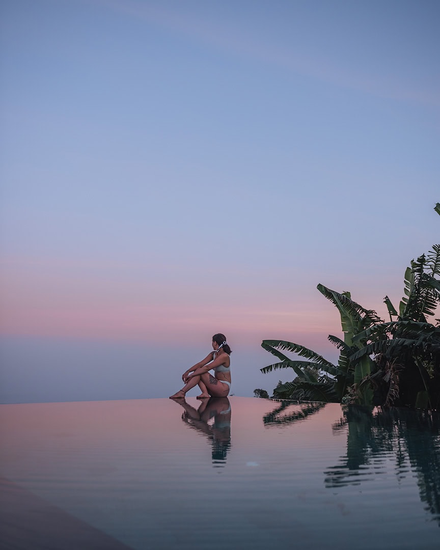 A woman sitting on the edge of a stunning infinity pool overlooking a pink and purple sunset sky.