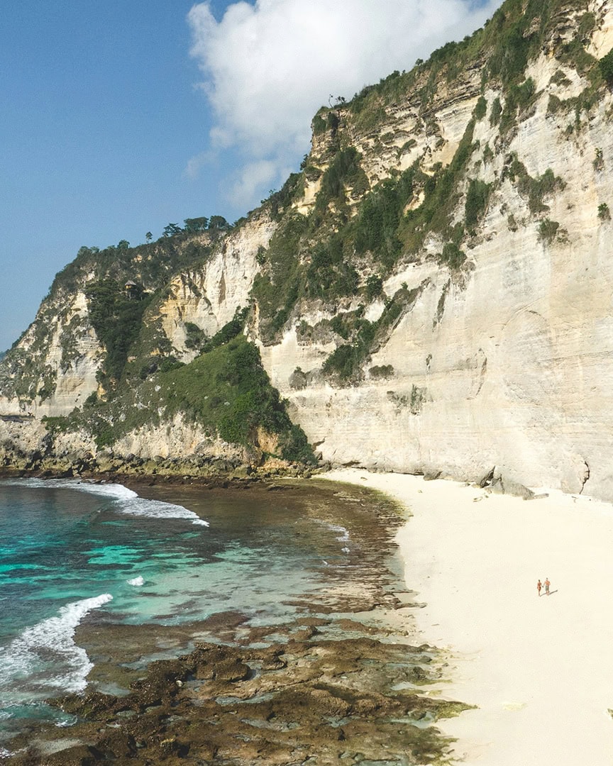 A secluded beach with white sand and turquoise water, bordered by high cliffs. Two people are holding hands walking along the beach. Diamond Beach Bali.