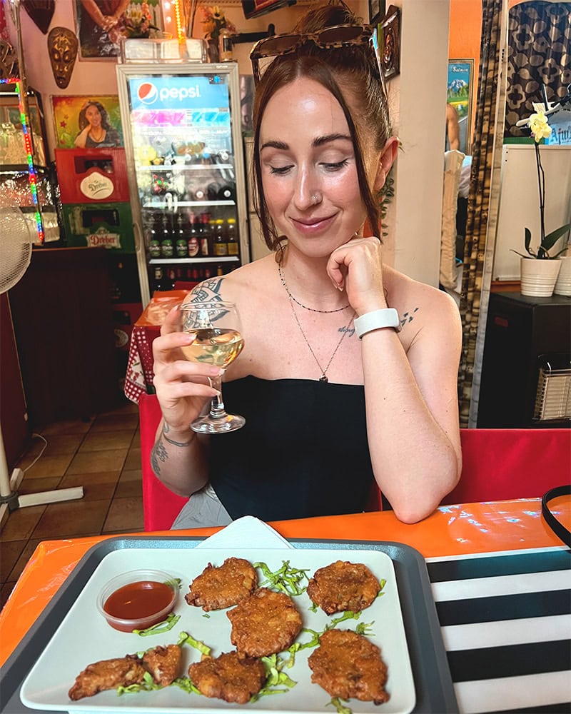 A woman seated at a table, holding a glass of white wine, with a plate of pakora in front of her.