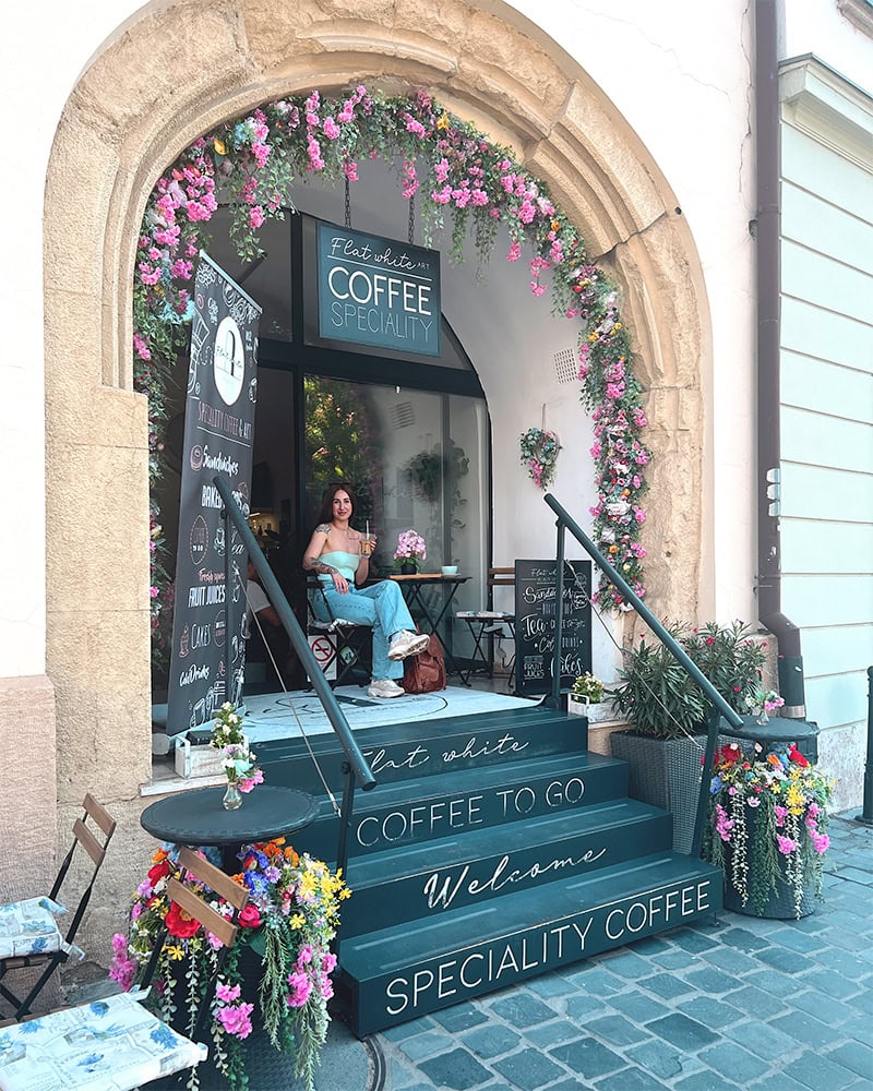 A woman seated outside a café in Budapest with pink flower decorations around the arched entrance.