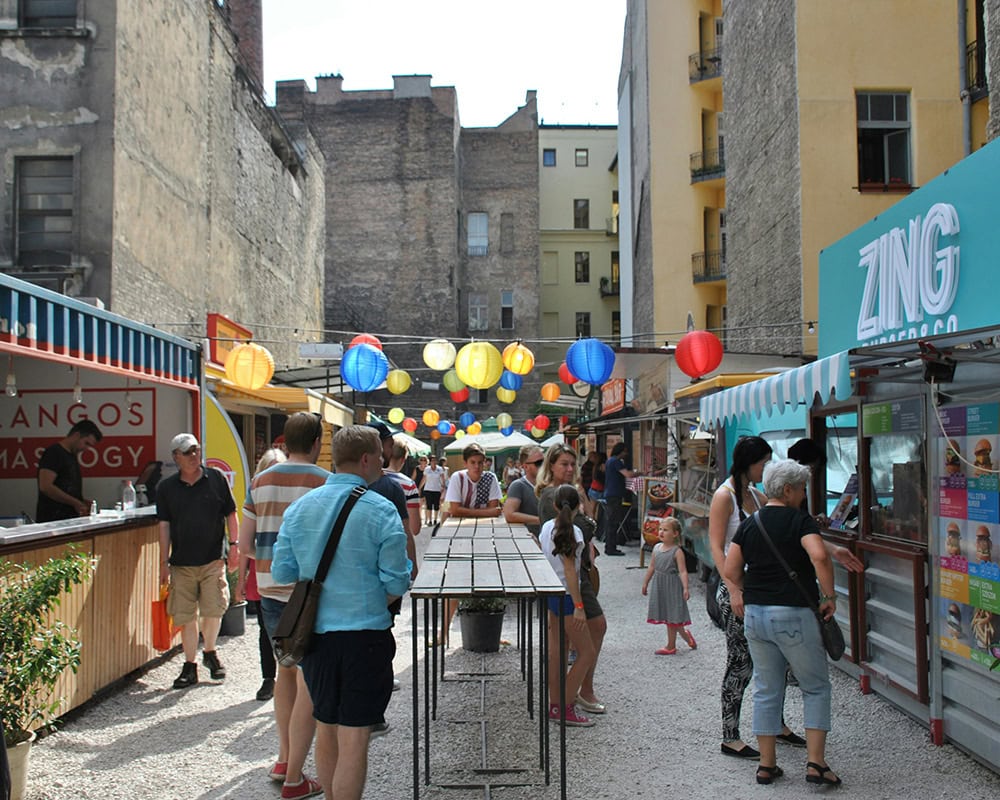 An outdoor food market with colorful lanterns hanging above, people browsing various food stalls. Street Food Karavan Budapest