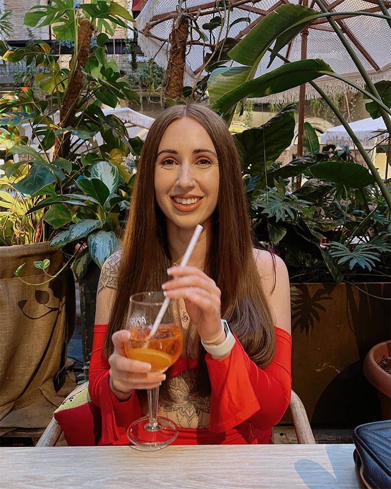 A woman smiling and holding a drink while seated at a table surrounded by lush plants. Twenty Six Budapest.