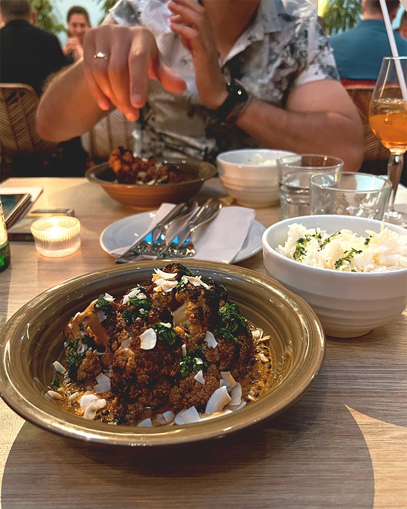 A close-up of a bowl of roasted cauliflower garnished with herbs, with rice and other dishes in the background.