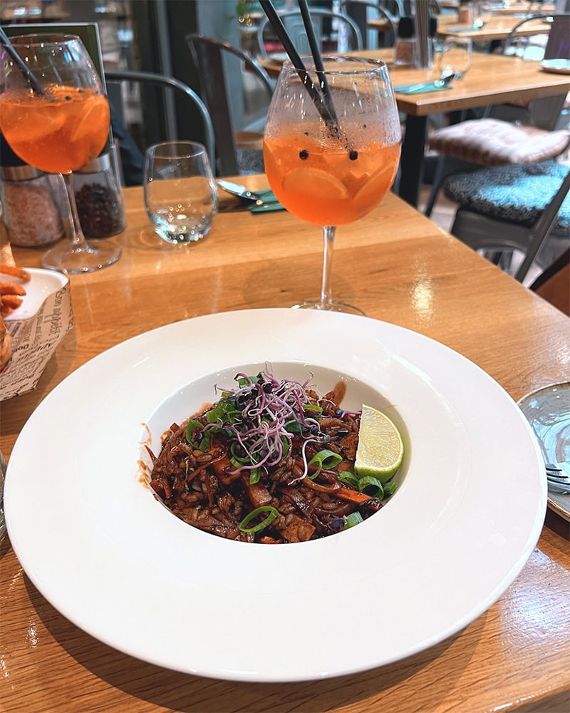 A plate of rice and vegetables garnished with green onions and lime, with two orange cocktails in the background. Vegan Garden Budapest