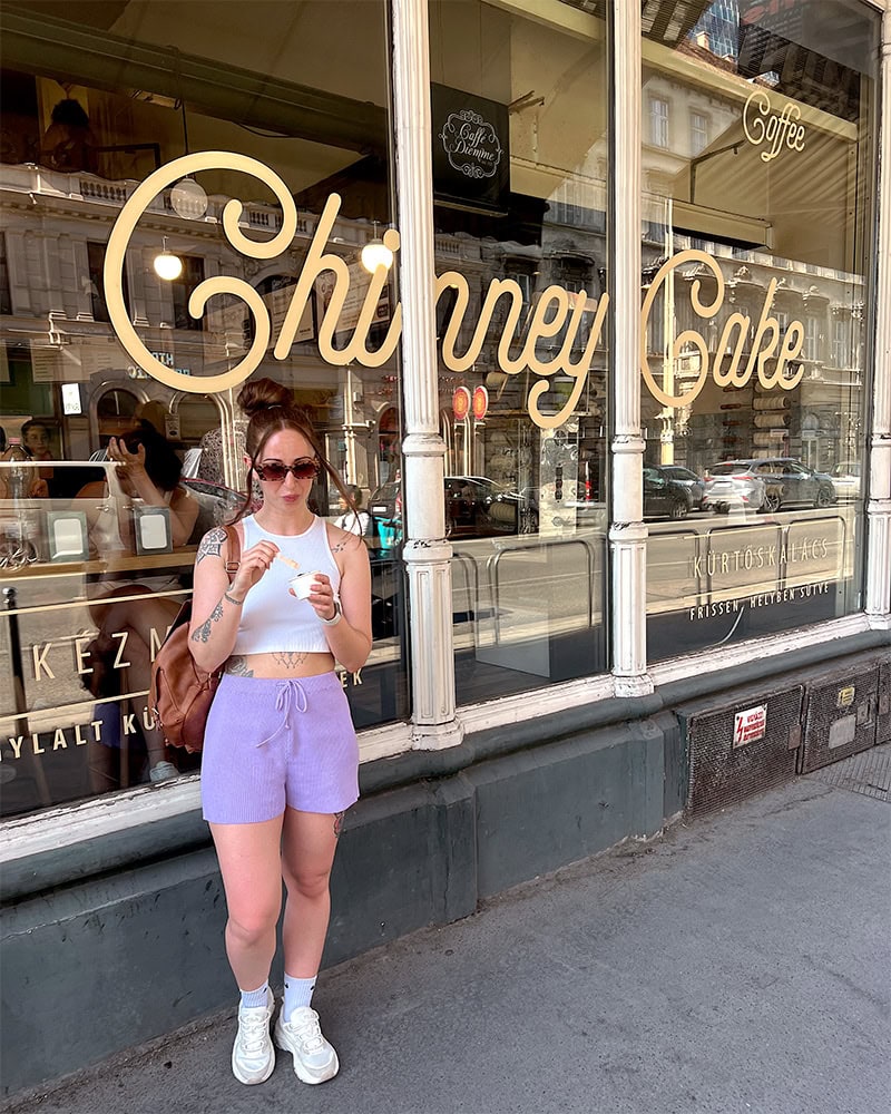A woman standing outside a café with "Chimney Cake," written across the window. She is holding and eating vegan ice cream. She is wearing a white crop top and lavender shorts, with sunglasses on her head.
